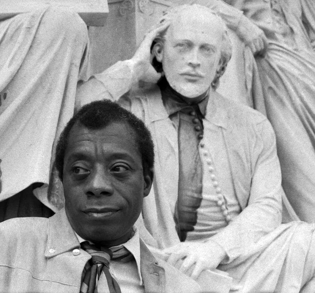 Portrait photograph of James Baldwin seated in front of a classical marble statue.