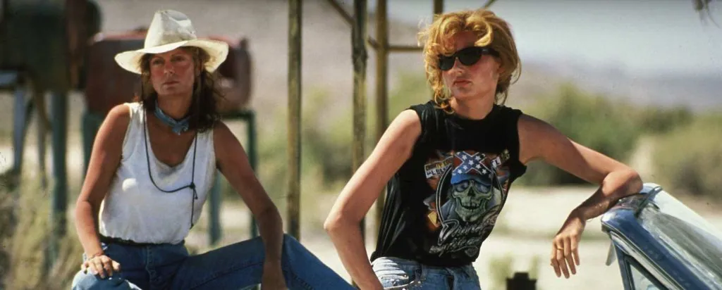 Film still from Thelma & Louise: the two women lean against their convertible in the desert, one in a straw hat, the other in sunglasses.