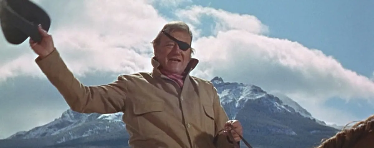 Film still from True Grit: John Wayne, wearing an eyepatch, raises his hat on horseback against a snowy mountain backdrop.
