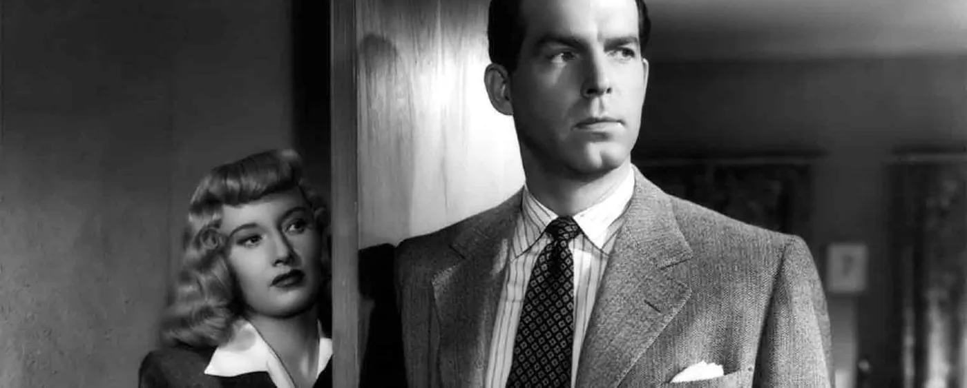 Film still from Double Indemnity: Barbara Stanwyck watches warily from a doorway behind Fred MacMurray in this black-and-white noir scene.