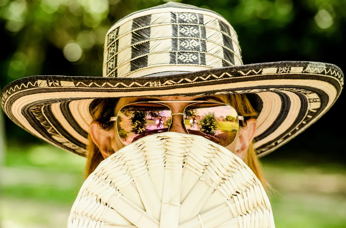 A woman in a wide-brimmed straw hat and sunglasses reflecting a sunlit landscape, holding a woven fan near her face.