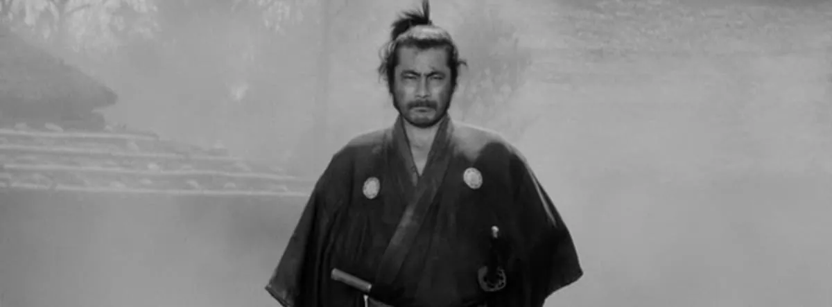 Film still from Yojimbo: the lone samurai, played by Toshiro Mifune, stands resolute in a misty village square.