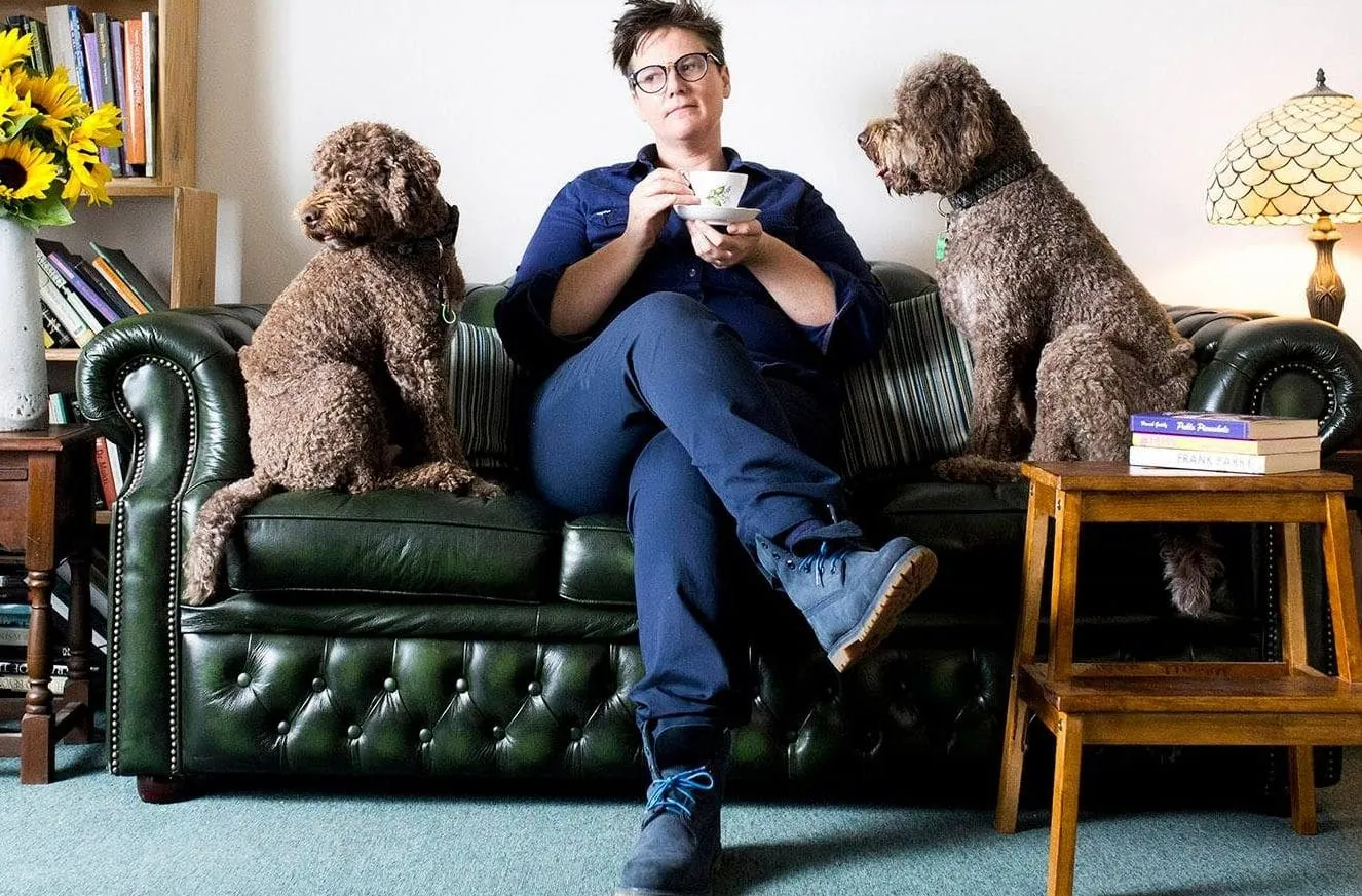 Hannah Gadsby sitting on a green leather couch with two dogs, holding a teacup.