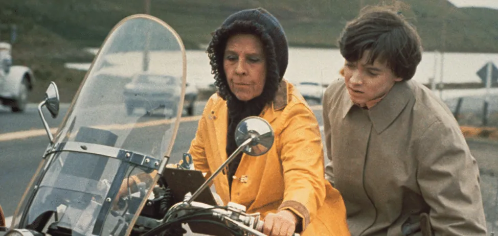Film still from Harold and Maude: Maude rides her motorcycle with Harold seated beside her.