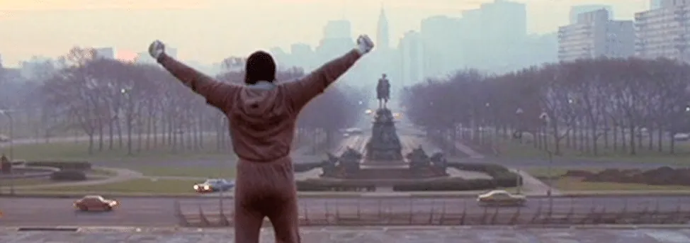 Film still from Rocky: Rocky stands with arms raised in triumph atop the Philadelphia Museum of Art steps, the city skyline behind him.