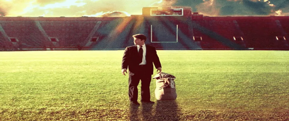 Film still from Rudy: a man in a suit stands alone on the football field holding a gear bag, the empty stadium bathed in golden light.
