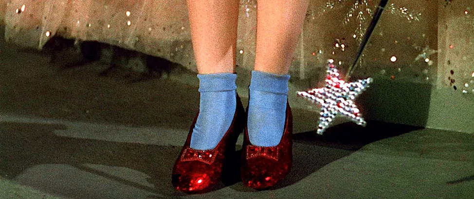 Film still from The Wizard of Oz: Dorothy's ruby slippers and blue socks, with the tip of Glinda's star wand beside them.