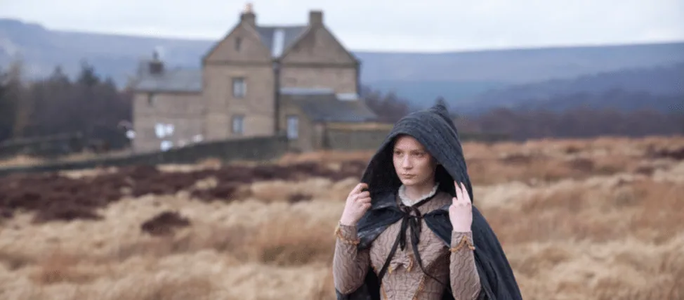 Film still from Jane Eyre: Jane stands on the windswept moor in a hooded cloak, the manor house visible in the distance.