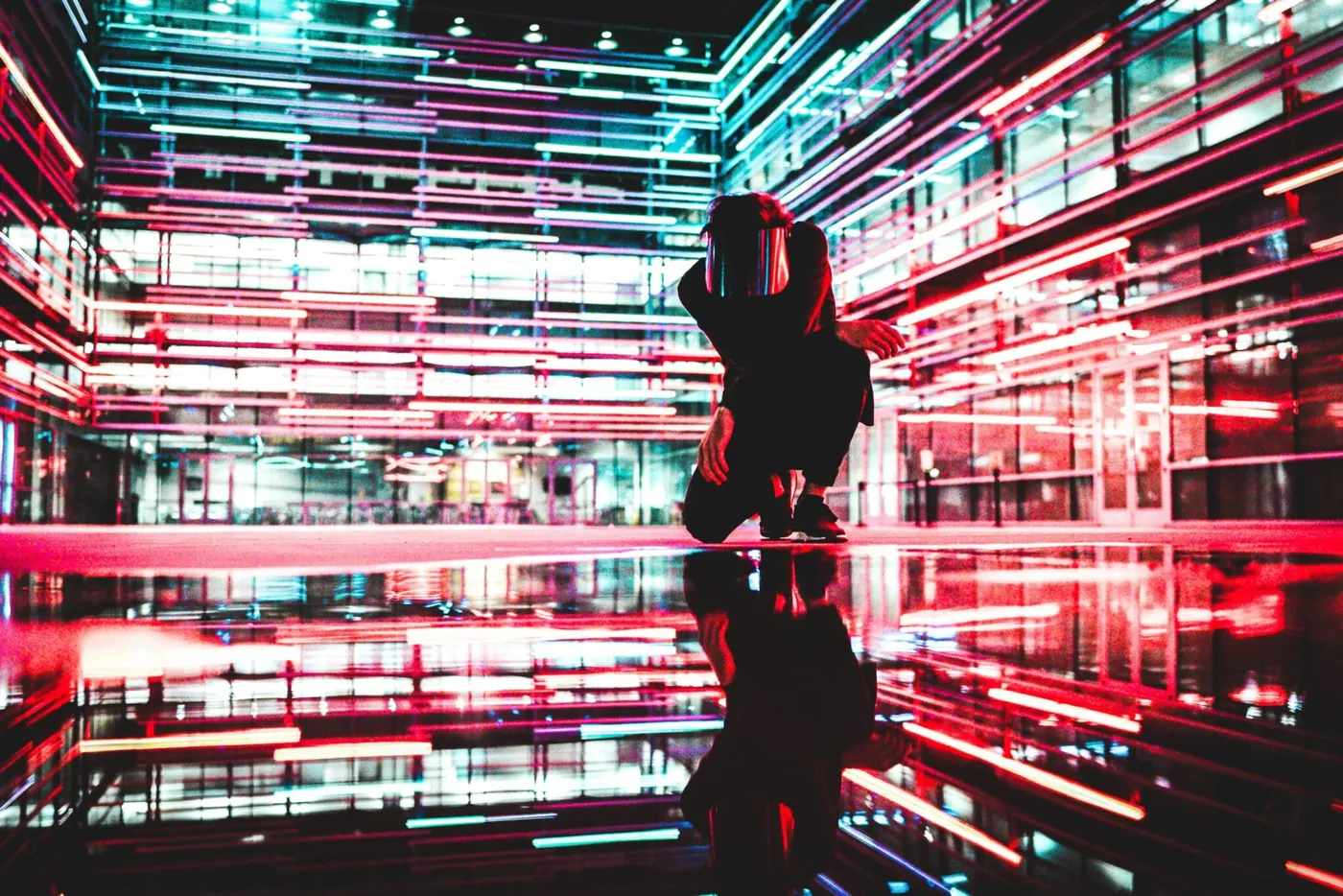 A person crouching low, reflected on wet ground amid streaking neon-lit architecture.