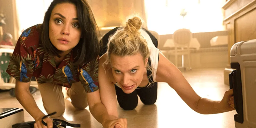 Film still from The Spy Who Dumped Me: the two friends crouch defensively behind furniture, looking alarmed.