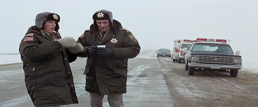 Film still from Fargo: two police officers in heavy winter coats stand on a snowy roadside holding coffee cups, patrol vehicles behind them.