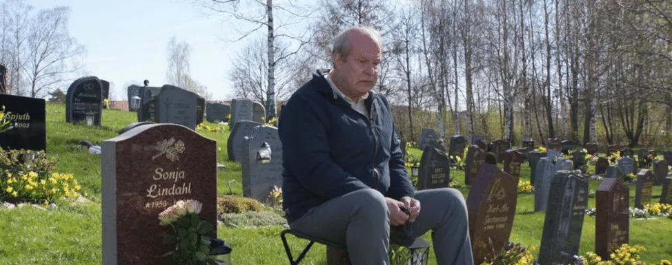 Film still from A Man Called Ove: Ove sits on a small stool beside his wife Sonja's grave in the cemetery.