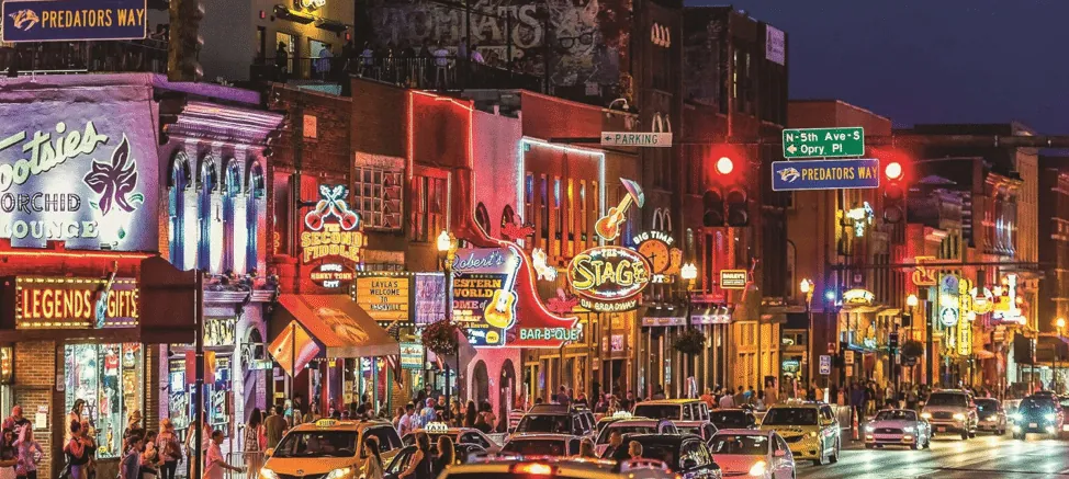 Nighttime view of Nashville's Broadway strip, lit up with neon honky-tonk signs and busy traffic.