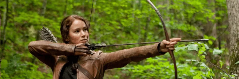 Film still from The Hunger Games: Katniss Everdeen draws her bow, aiming through the forest.