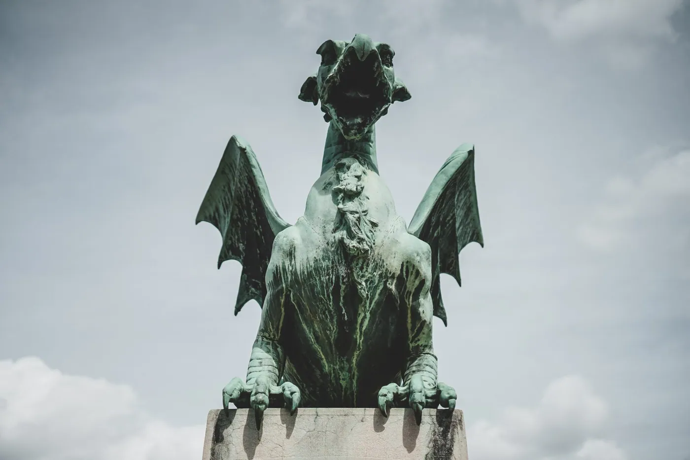 A weathered bronze dragon statue with spread wings perched on a stone pedestal against a cloudy sky.