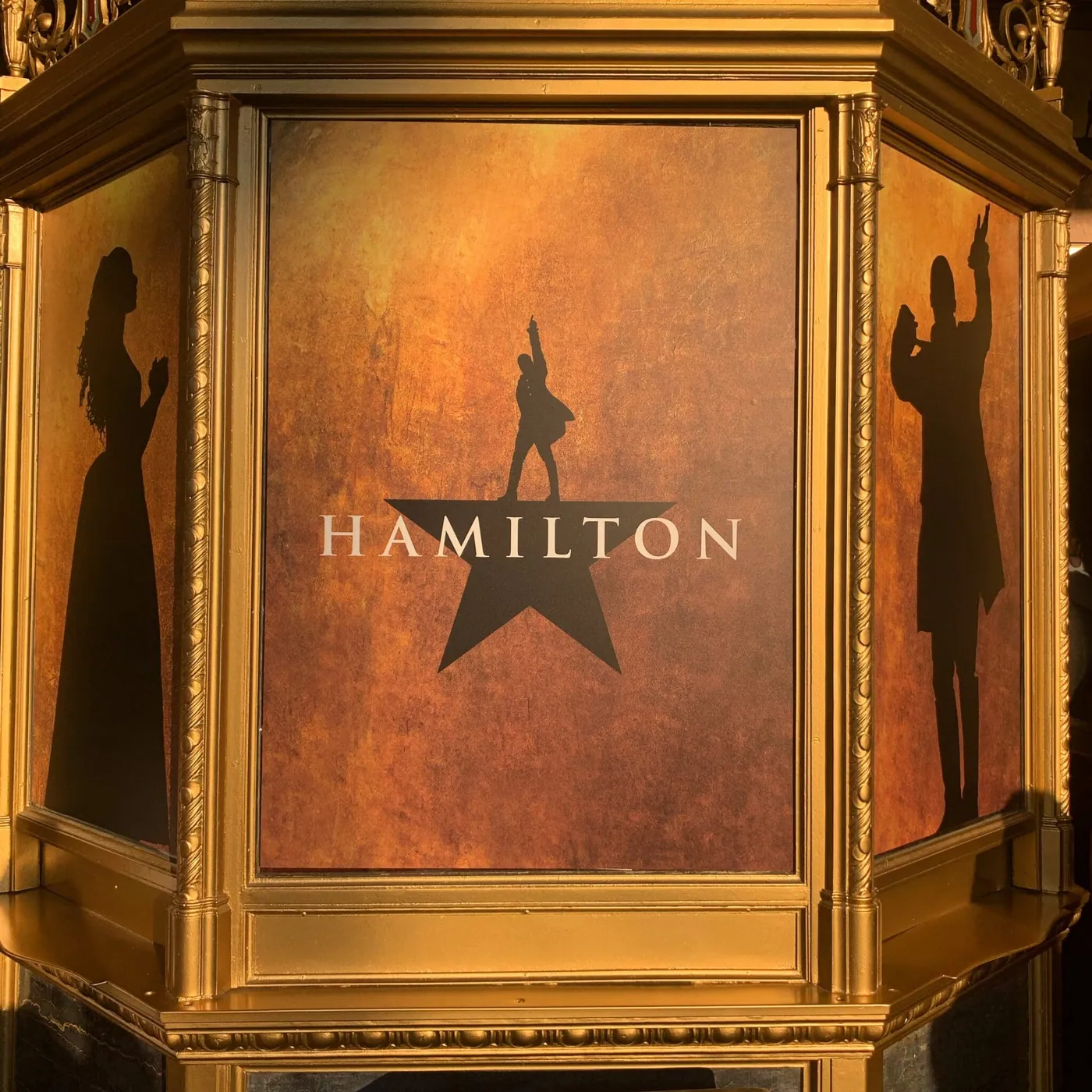 A gold theater lobby display for the musical Hamilton, showing the show's star logo flanked by silhouettes of two characters.