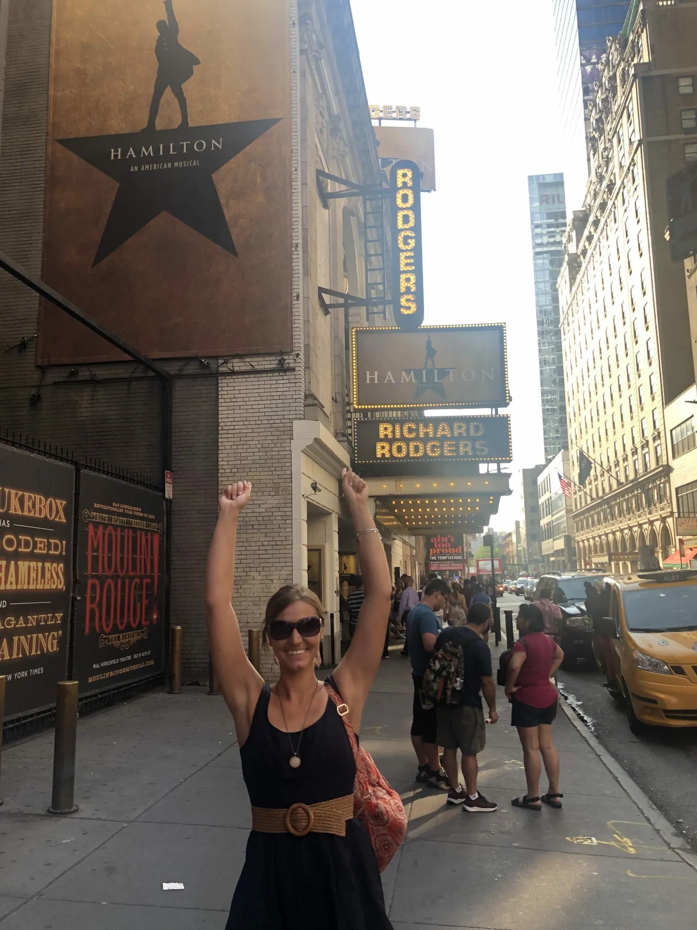 A woman raises her arms in celebration on a New York City sidewalk outside the Richard Rodgers Theatre, its marquee reading Hamilton.