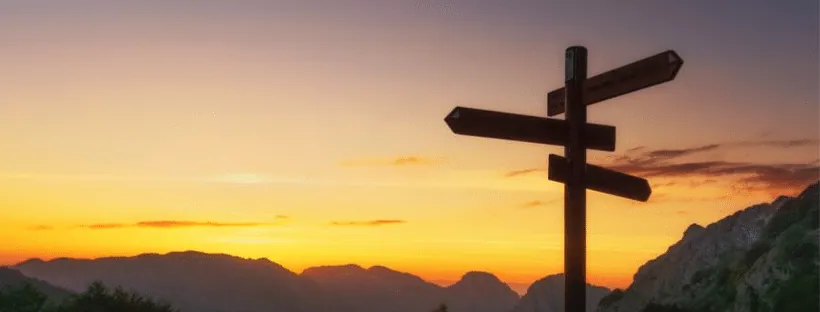 A wooden signpost with four blank direction arms, silhouetted against an orange sunset over distant mountains.