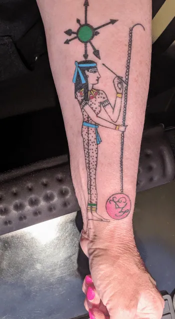 A tattoo on a forearm depicting an ancient Egyptian figure in profile, holding a knotted cord attached to a pink pendant, beneath a sunburst symbol.