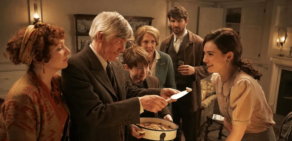 Film still from The Guernsey Literary and Potato Peel Pie Society: the islanders gather around a homemade potato peel pie as an elderly man presents it to Juliet.