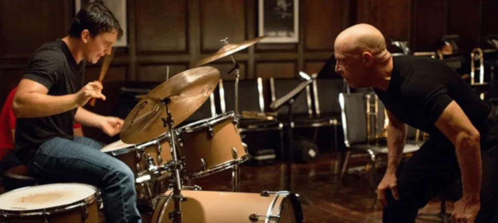 Film still from Whiplash: Andrew plays drums intensely while his conductor, Fletcher, leans in and glares at him.