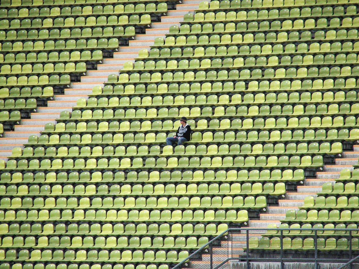 A single person sitting alone amid many rows of empty green stadium seats.