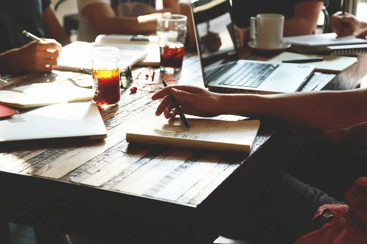 People gathered around a wooden table with laptops, notebooks, and drinks, writing and discussing.
