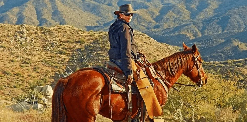 A woman in a cowboy hat and sunglasses sits on a horse overlooking a desert mountain landscape.