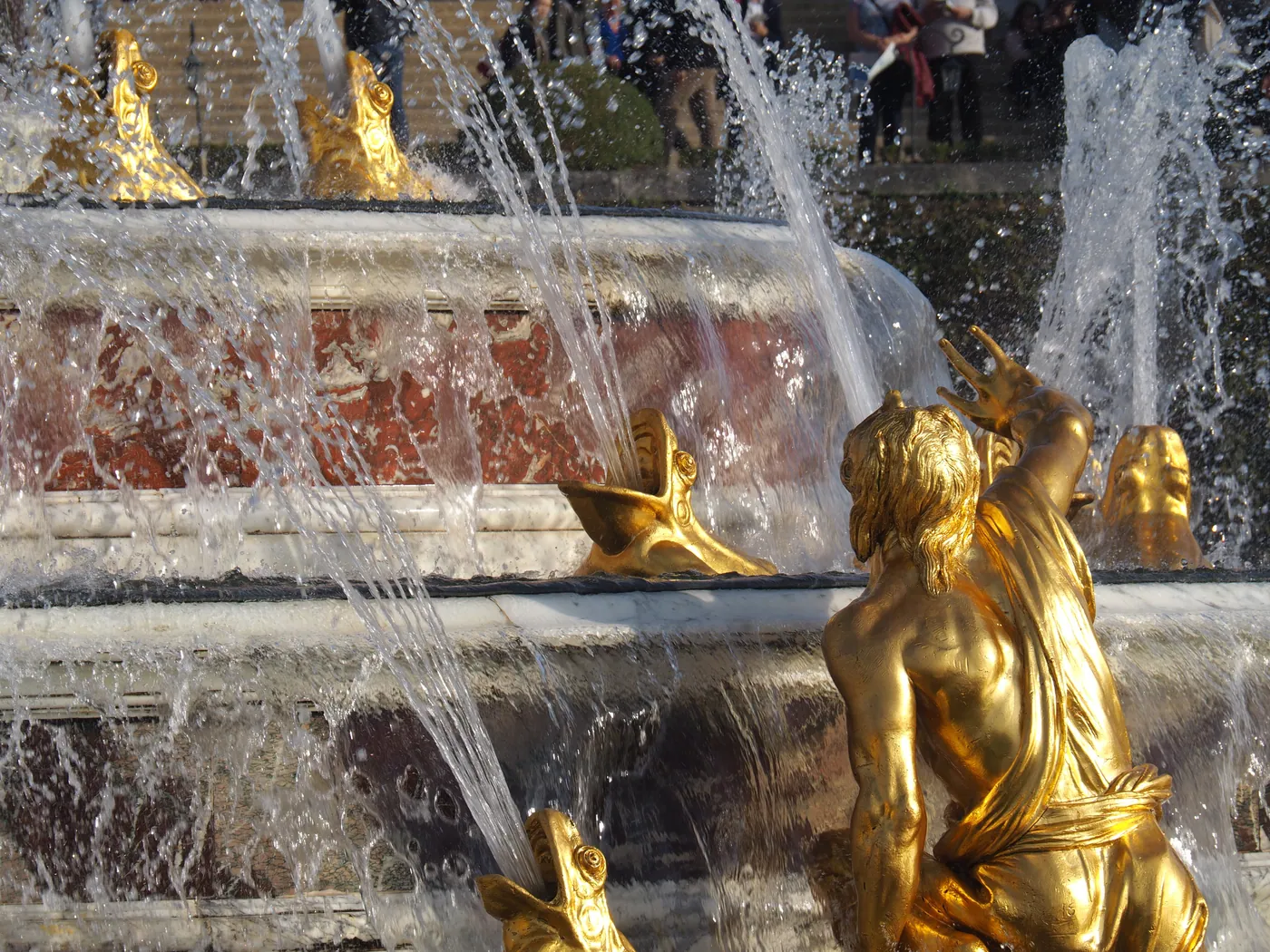 A gilded bronze statue and dolphin-head fountains spraying water, part of an ornate palace garden fountain.