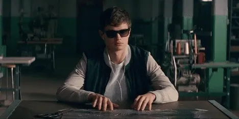 Film still from Baby Driver: the getaway driver in sunglasses and earbuds seated at a table.