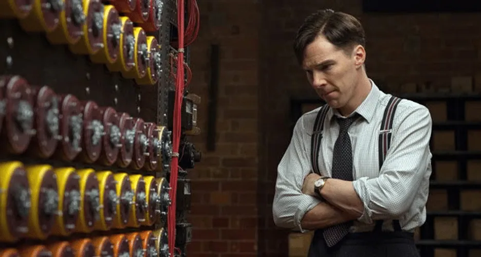 Film still from The Imitation Game: Alan Turing, arms crossed, stands beside the rows of rotors on his codebreaking machine.