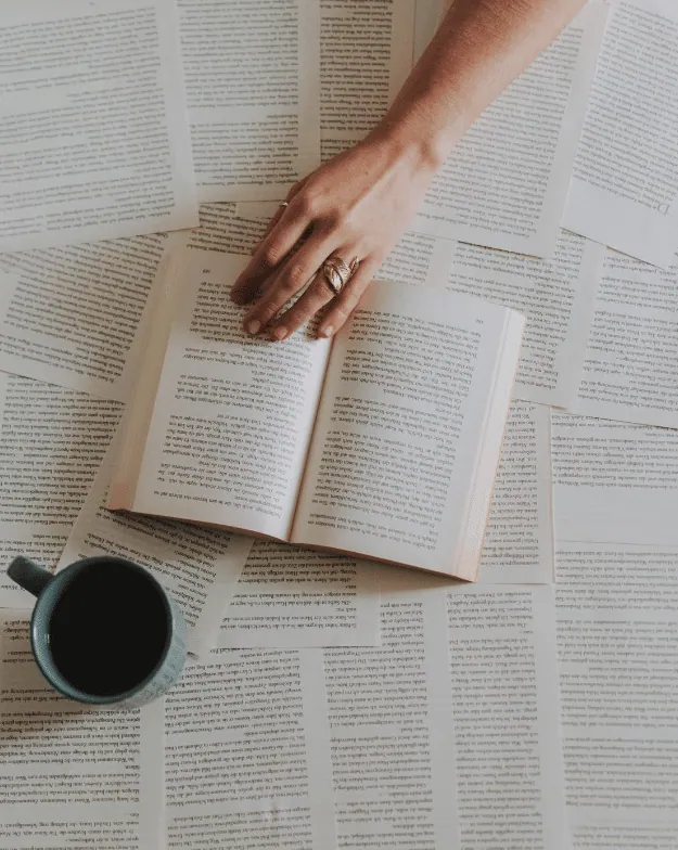 An open book on a table scattered with book pages, a hand with a ring reaching toward it, and a mug of coffee nearby.