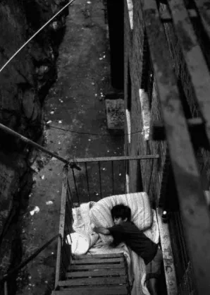A person sleeping on a mattress on a fire escape above a grimy alley, in black and white.