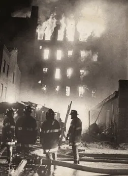 A black-and-white photograph of New York in the 1970s. Firefighters stand silhouetted in the foreground while flames pour from the windows of every floor of a burning multi-storey tenement behind them.