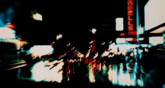 A blurred night-time view through a taxi windscreen. Red, white and green neon smears across the wet glass, with an illuminated hotel sign just legible at the right.