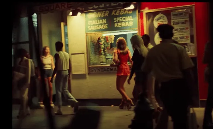 A night-time city street outside a lit counter advertising doner kebab, Italian sausage and special kebab, with a partly hidden sign reading Square above it. Women in short dresses stand and walk along the pavement among other passers-by under red and yellow shop light.