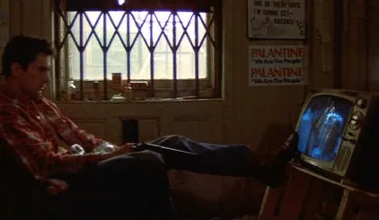 Travis pushing his boot against the television screen, about to knock it over, Palantine campaign posters on the wall behind him.