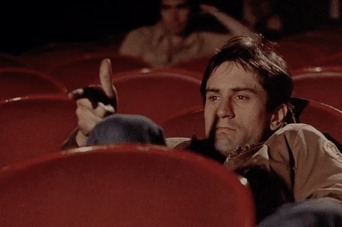 Travis Bickle sitting alone in a movie theater, pointing his hand like a gun at the screen.