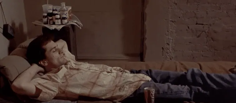 Travis Bickle lying on his bed beside a nightstand of pill bottles in his sparse apartment.