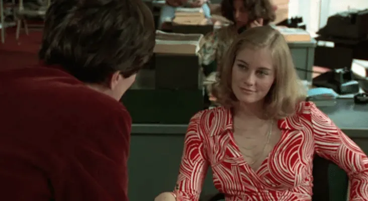 The Lovers Meet scene from Taxi Driver. Travis, seen from behind in his wine-coloured jacket, leans across a desk in the Palantine campaign office towards Betsy, who looks back at him in a red and white patterned dress. Typewriters and stacked paperwork fill the office behind her.