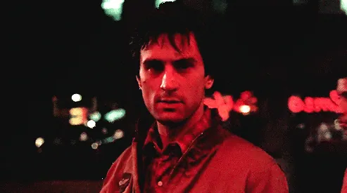 Travis Bickle, lit in red, standing alone on a neon-lit street at night.