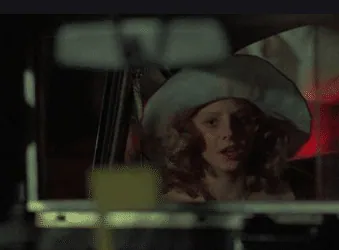 A young girl in a floppy hat getting into Travis's taxi at night.