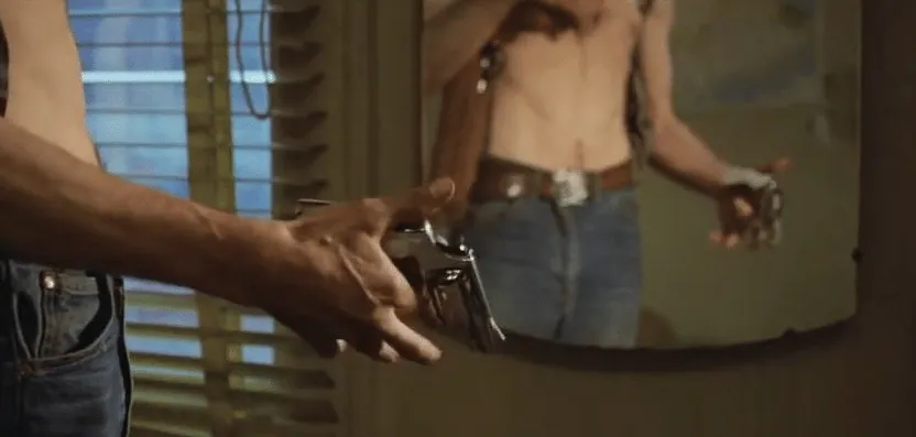 A shirtless Travis Bickle practicing drawing a handgun with a spring-loaded wrist holster in front of a mirror.