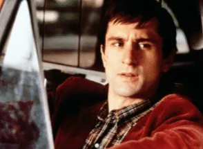 Robert De Niro as Travis Bickle behind the wheel of his taxi, glancing toward the camera.