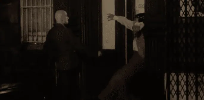 Sport falling backward against a doorway after being shot, in dim, blood-toned lighting.