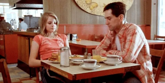 Travis and Iris sitting tensely across from each other at a diner breakfast table.