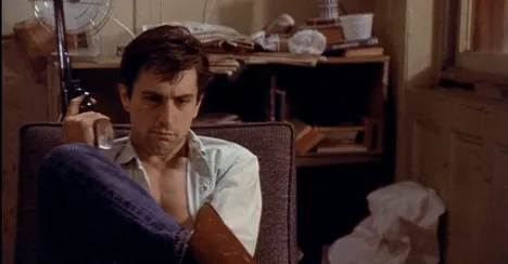 Travis sitting slumped in a chair holding a gun, looking despondent in his cluttered apartment.