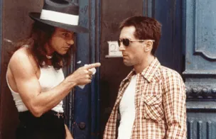 Sport, in a wide-brimmed hat, pointing at Travis on the street outside a doorway.