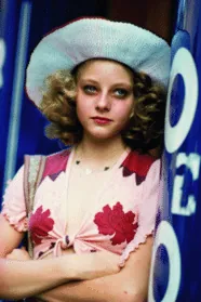 Iris standing with arms crossed against a doorway, in her floppy hat and floral top.