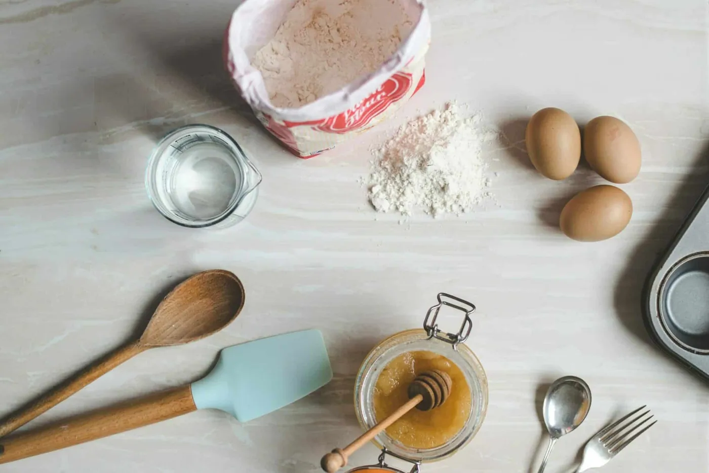 Flat-lay of baking ingredients and utensils: flour, eggs, a honey jar, and a measuring cup of water. (Image by Wooden Earth Cutlery)